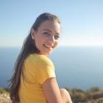 Young woman in a yellow shirt smiles by the sea, enjoying the outdoor view.